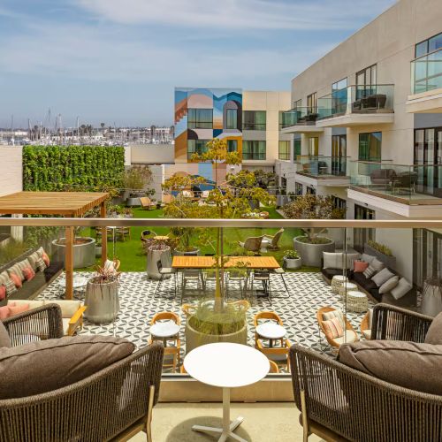 A sunny resort poolside scene with lounge chairs, umbrellas, and a dining table set, a modern building in the background, and blue sky.