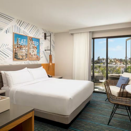 A bright hotel room with a large bed, cozy seating, a desk, and a flat-screen TV, plus warm lighting and a balcony with city views.