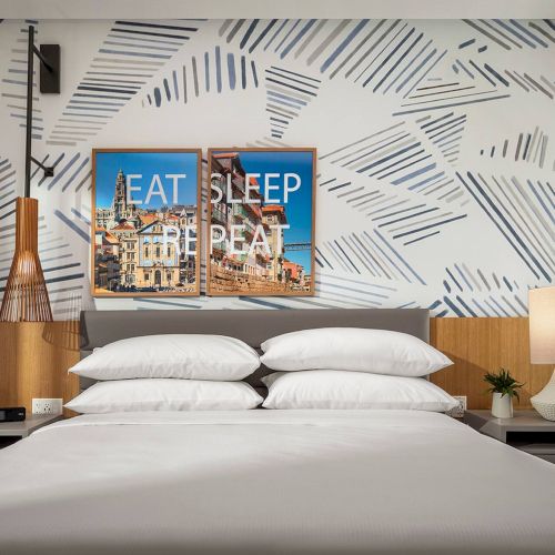 A bright bedroom with two beds, a shared nightstand, large window, and a colorful wall art piece above the headboards, creating a cozy, modern vibe.