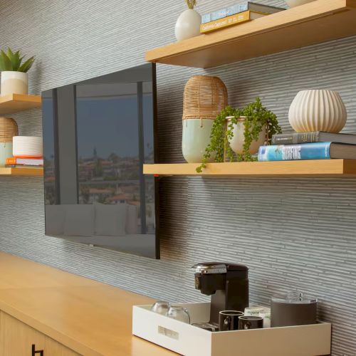 A modern living room with a wall-mounted TV, wooden shelves, a cozy sofa, and decorative items on a light background.