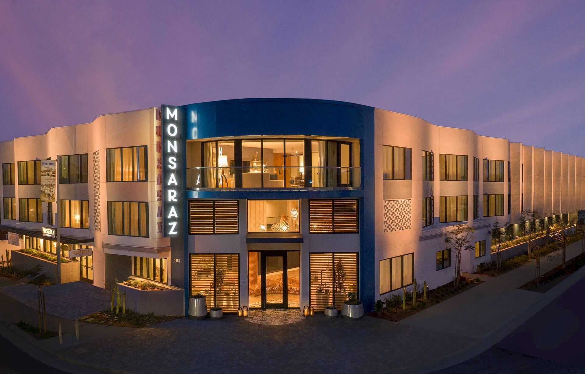 A modern, multi-story office building with a curved facade and a prominent glowing &ldquo;MONSARAZ&rdquo; sign, illuminated at dusk.