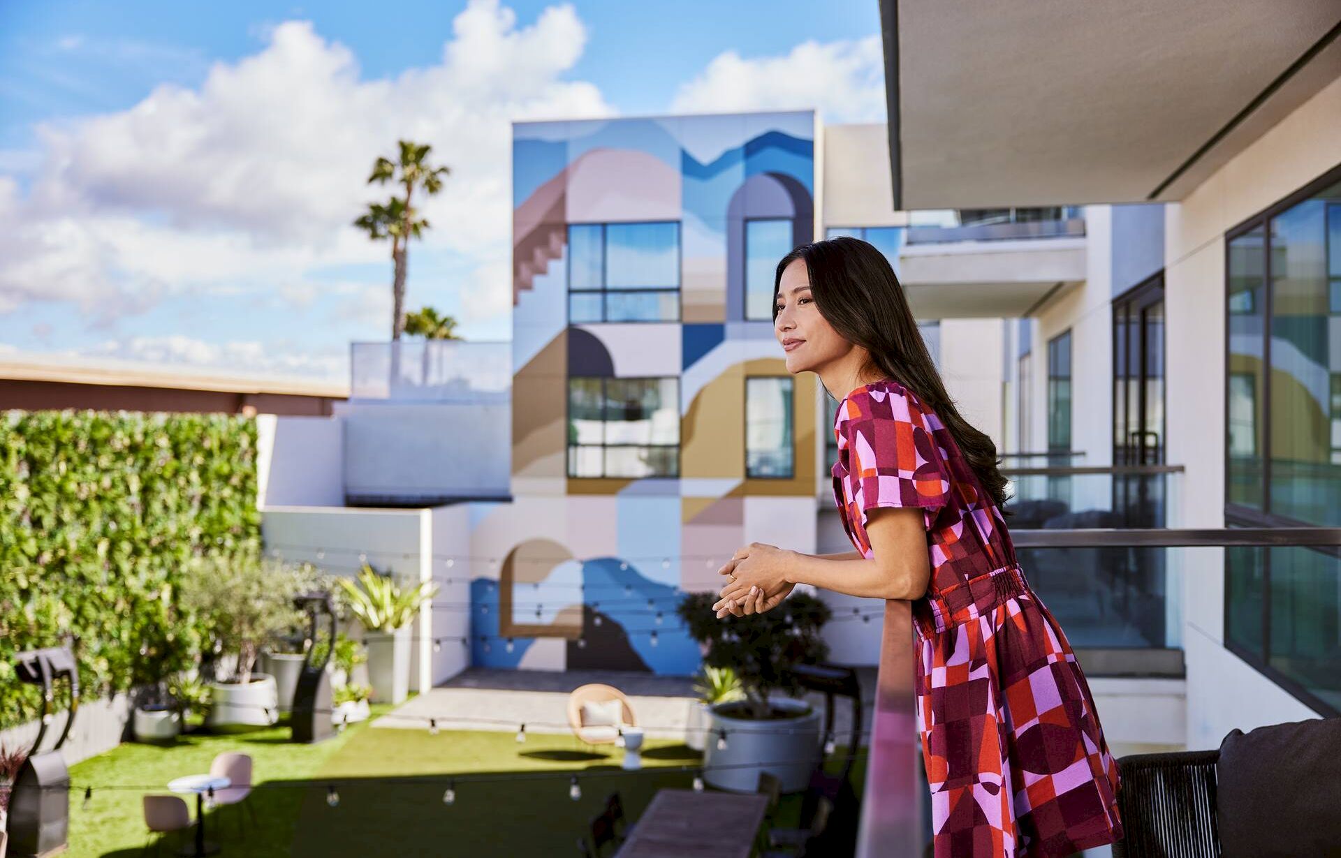 A woman in a red patterned dress stands on a balcony overlooking a colorful geometric mural and modern building, sunny day, palm trees nearby.