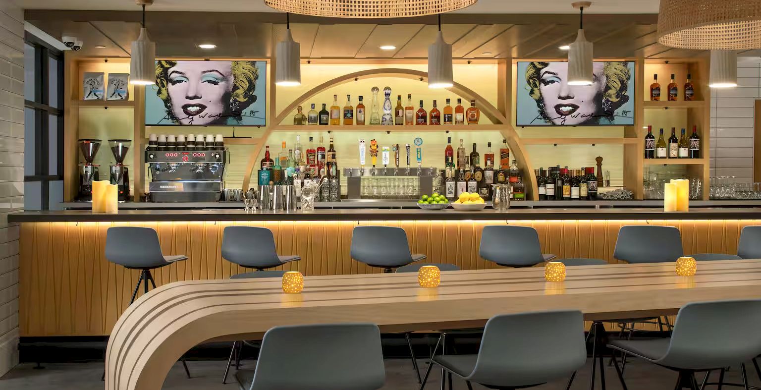 A stylish bar with a curved wooden counter, bar stools, shelves of bottles, two Marilyn Monroe portraits, and warm pendant lighting.